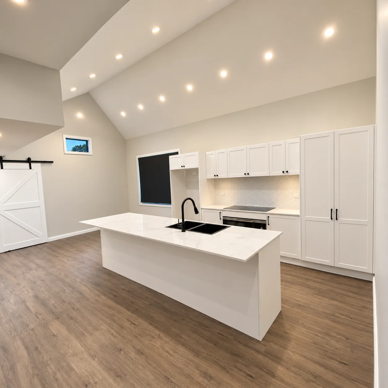 A white kitchen with a stone island, black tapware and a high ceiling with recessed lights.